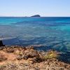 Scogliera rocciosa con mare cristallino, tonalità blu e verde, isola in lontananza.