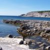 Paesaggio costiero roccioso con acqua limpida, promontorio in lontananza.