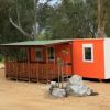 Una piccola casa prefabbricata arancione con veranda di legno e tetto verde in un'area alberata.