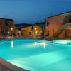 Piscina accesa di sera, con ombrelloni intorno e edifici sullo sfondo.