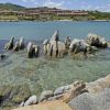 Scogli affioranti in mare cristallino, complesso residenziale sullo sfondo.