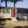 Casa mobile beige con tende blu, porta rossa, patio con tavolo bianco e sedie, sotto alberi.